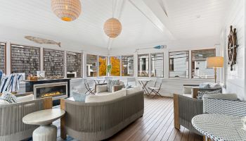 The image shows a cozy sunroom with wooden floors, stylish furniture, a fireplace, and large windows providing a bright, inviting atmosphere.