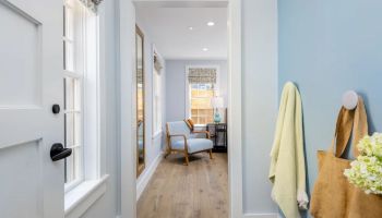 A hallway with light blue walls contains a beige bag and yellow towel hanging on hooks; it leads to a room with a chair, lamp, and large windows.