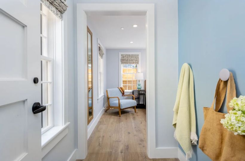 A hallway with light blue walls contains a beige bag and yellow towel hanging on hooks; it leads to a room with a chair, lamp, and large windows.