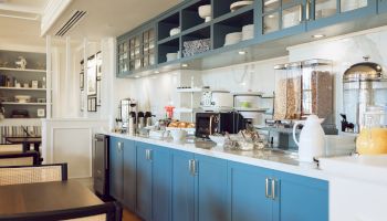 A breakfast buffet setup with cereals, pastries, drinks, and kitchenware on blue cabinets, in a cozy dining area setting.