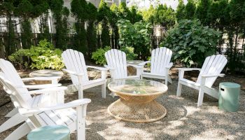A sunny backyard seating area with white Adirondack chairs around a woven patio table, surrounded by lush green plants and shrubs.