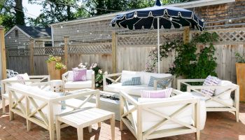 A sunny backyard patio with white lounge furniture, floral pillows, and string lights under a navy canopy, perfect for relaxing.