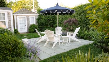 A backyard patio with white Adirondack chairs and a dark striped cantilever umbrella above the garden.