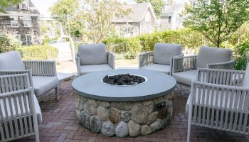 A backyard patio with cushioned lounge chairs around a stone fire pit filled with black rocks.