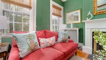 A cozy living room with a red sofa adorned with pineapple-themed cushions, green walls, a white fireplace, and large windows.