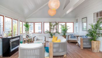 A cozy sunroom with large windows, two round armchairs, a fireplace, indoor plants, and pendant lights, creating a bright and airy ambiance.