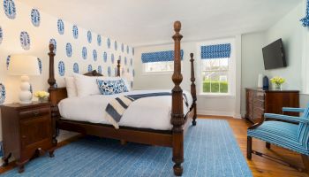 A bedroom with blue and white decor, featuring a four-poster bed, armchair, dresser, TV, and windows with patterned shades.