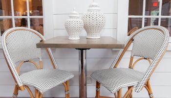 The image shows a small table with two wicker chairs, featuring white cushions with a dot pattern and two decorative white jars on the table.