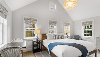 A cozy bedroom with a large bed, blue throw, multiple windows, wicker chairs, lamps, and a modern light fixture on the ceiling.