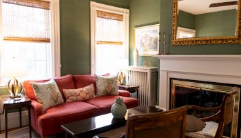 A cozy living room with a red sofa, patterned cushions, green walls, a fireplace with a large mirror, and wooden chairs by the table.