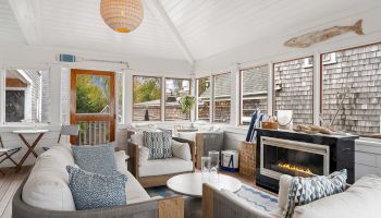 A cozy sunroom with white sofas, a fireplace, and nautical decor, including an oar and fish sculpture. Large windows offer outdoor views.