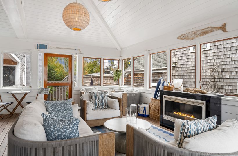 A cozy sunroom with white sofas, a fireplace, and nautical decor, including an oar and fish sculpture. Large windows offer outdoor views.