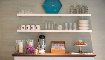 A coffee station with cups, mugs, glasses, plates, a coffee dispenser, creamers, flowers, and pastries displayed on a countertop and shelves.