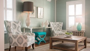 A cozy living room with patterned armchairs, a wooden coffee table, table lamps, and decorative vases. Light green walls and an area rug complete the setup.