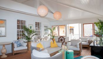 A cozy, modern living space with comfortable seating, potted plants, warm lighting, and a large wooden ship wheel as a decorative element in the corner.