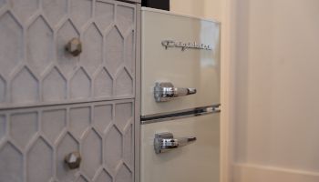 The image shows a retro-style refrigerator with chrome handles next to a textured cabinet, creating a vintage kitchen aesthetic.