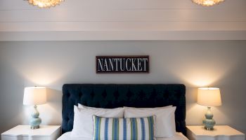 A stylish bedroom features a navy tufted headboard, striped cushion, "NANTUCKET" wall sign, two lamps, and decorative lighting.