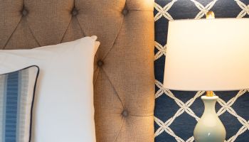 A bed with a tufted headboard, a striped pillow, a bedside lamp, and a rattan nightstand, set against a rope-patterned wallpaper.