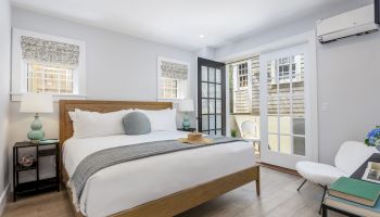 A modern bedroom with a wooden bed, white bedding, bedside tables, and a chair by French doors leading to a small patio area with two chairs.