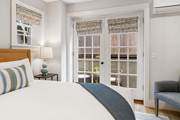 A cozy bedroom features a bed with striped pillows, a bedside lamp, and French doors leading to a patio with chairs and plants.