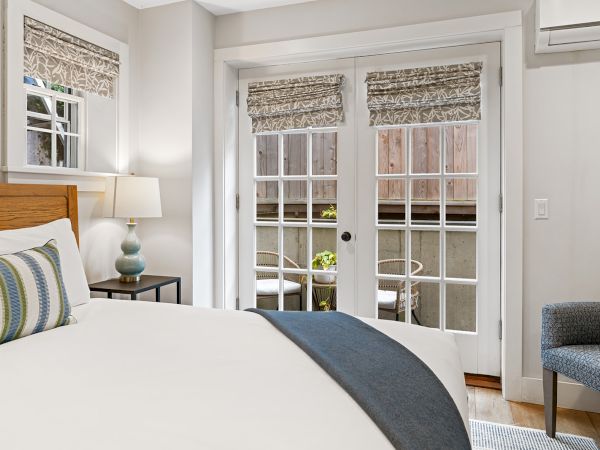 A cozy bedroom features a bed with striped pillows, a bedside lamp, and French doors leading to a patio with chairs and plants.