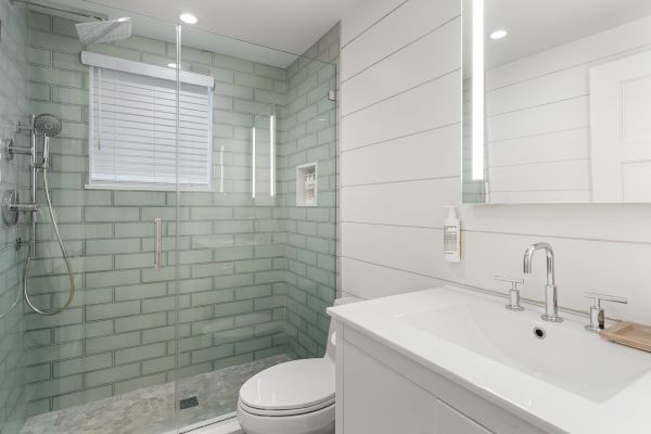 The image shows a modern bathroom with a glass shower, green tiles, a toilet, and a sleek vanity with a mirror and illuminated lighting.