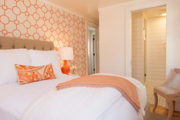 A cozy bedroom with an orange and white design, featuring a bed, patterned wallpaper, a nightstand with a lamp, a chair, and a doorway to a bathroom.