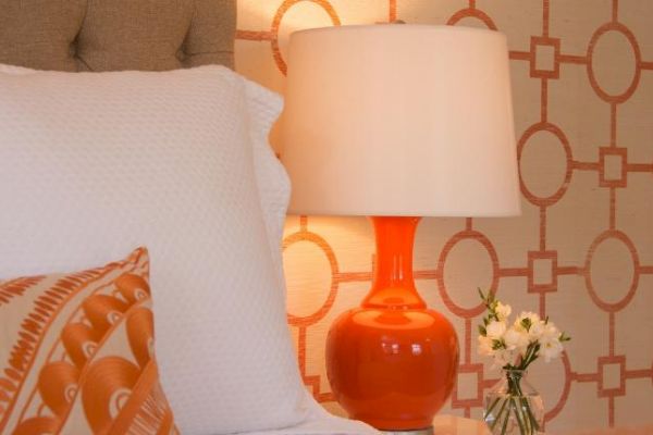 A cozy bedroom scene featuring an orange lamp, white flowers in a vase, a patterned pillow, and a stylish geometric wallpaper design.