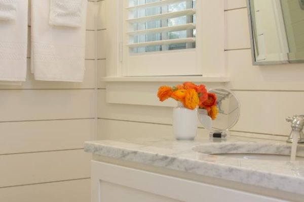 A bathroom with white wood paneling, a marble countertop, and a small vase of orange flowers by the sink, with towels hanging on the wall.