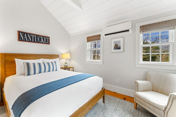 A cozy bedroom with a bed, striped pillow, blue throw, armchair, two windows, wall art, and a "Nantucket" sign above the bed.