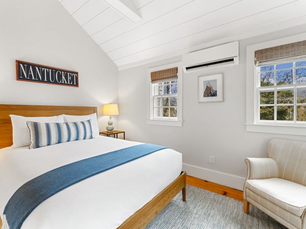A cozy bedroom with a bed, striped pillow, blue throw, armchair, two windows, wall art, and a "Nantucket" sign above the bed.