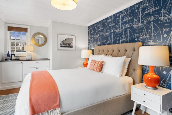 A bedroom with a nautical theme features a large bed, orange lamps, blueprint wallpaper, and decor elements like a mirror and framed picture.