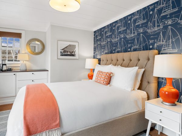 A bedroom with a nautical theme features a large bed, orange lamps, blueprint wallpaper, and decor elements like a mirror and framed picture.