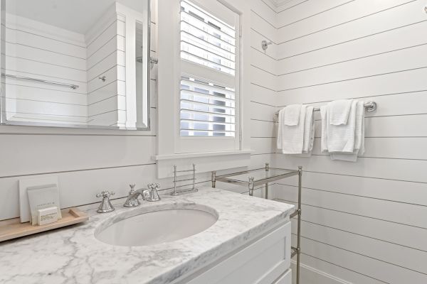 A bathroom with white shiplap walls, a marble countertop, a mirror, towels on a rack, and plantation shutters on the window.