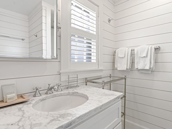 A bathroom with white shiplap walls, a marble countertop, a mirror, towels on a rack, and plantation shutters on the window.