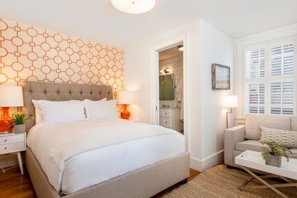 A cozy bedroom with a patterned accent wall, a large bed, two lamps on nightstands, a sofa, a coffee table, and a bathroom in the background.