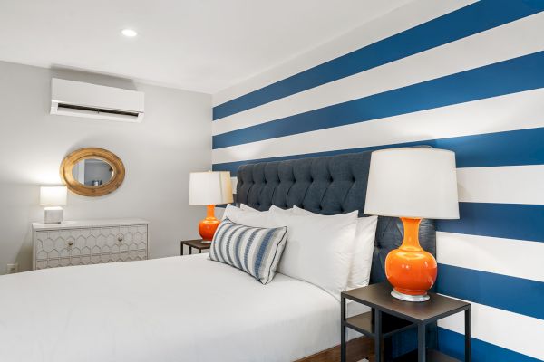 A modern bedroom features a striped blue and white accent wall, tufted headboard, orange lamps, and a wooden framed mirror on a dresser.