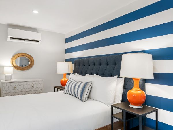A modern bedroom features a striped blue and white accent wall, tufted headboard, orange lamps, and a wooden framed mirror on a dresser.