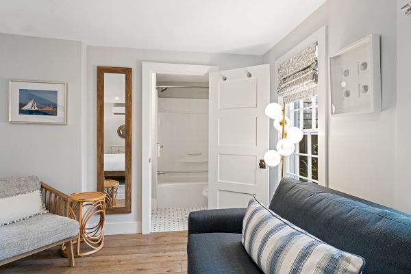 A cozy room with a sofa, patterned pillow, floor lamp, mirror, and a view into a bathroom. Artwork and window blinds complete the decor.