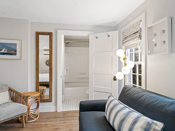 A cozy room with a sofa, patterned pillow, floor lamp, mirror, and a view into a bathroom. Artwork and window blinds complete the decor.