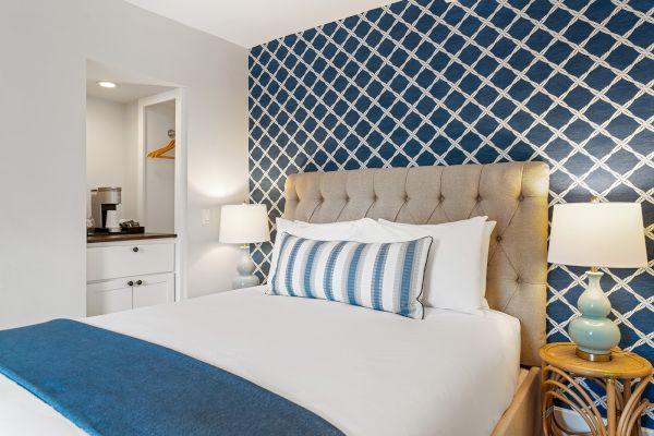 A cozy bedroom with a beige headboard, blue patterned wall, striped pillow, side table, lamp, and small nook with a coffee maker.