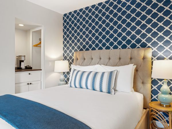 A cozy bedroom with a beige headboard, blue patterned wall, striped pillow, side table, lamp, and small nook with a coffee maker.