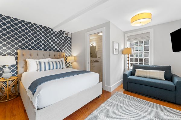 A cozy bedroom with a tufted bed, blue sofa, patterned wallpaper, lamps, and a small bathroom. A rug covers the wooden floor.