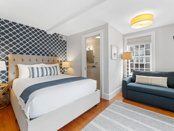 A cozy bedroom with a tufted bed, blue sofa, patterned wallpaper, lamps, and a small bathroom. A rug covers the wooden floor.