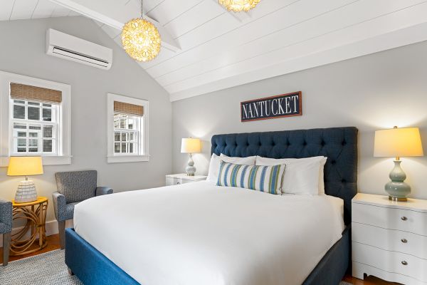 A cozy bedroom with a bed, blue tufted headboard, lamps, armchair, and "Nantucket" sign on the wall, under a vaulted ceiling.