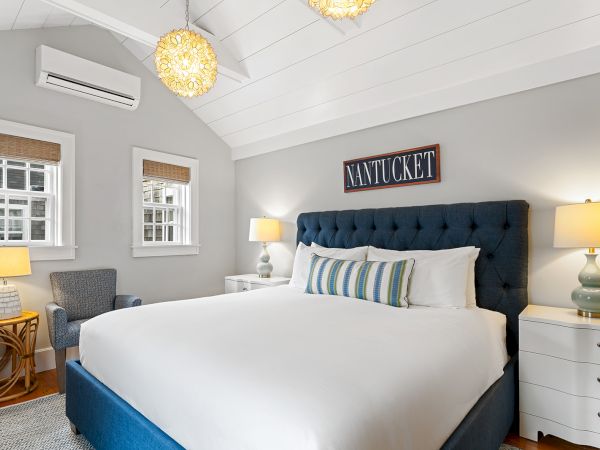 A cozy bedroom with a bed, blue tufted headboard, lamps, armchair, and "Nantucket" sign on the wall, under a vaulted ceiling.