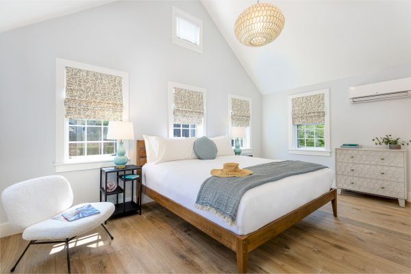 This image shows a modern, bright bedroom with a bed, white walls, wooden floor, a chair, and a dresser, featuring natural light and decor.