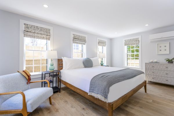 A bright bedroom with a large bed, light gray walls, wooden floor, an armchair, lamps on side tables, a dresser, and multiple windows with blinds.