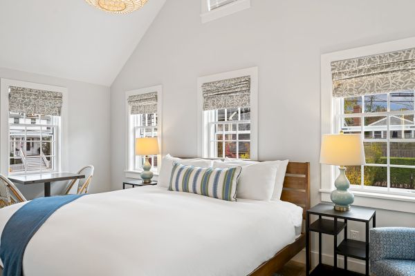 A cozy bedroom with a large bed, striped pillow, a blue throw, side tables with lamps, and windows with patterned blinds.