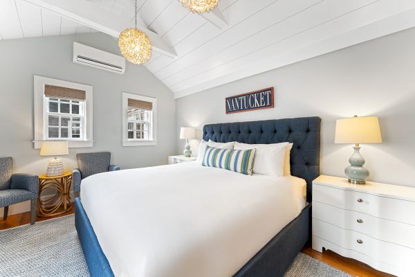 A cozy bedroom with a large bed, blue headboard, lamps, chairs, and a "Nantucket" sign on the wall.